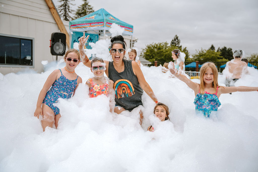 Group of children and an adult having fun in a foam party outdoors. The kids are smiling and wearing colorful swimsuits, while the adult in sunglasses and a tank top joins in the excitement. The background shows a party setup with a canopy and foam.