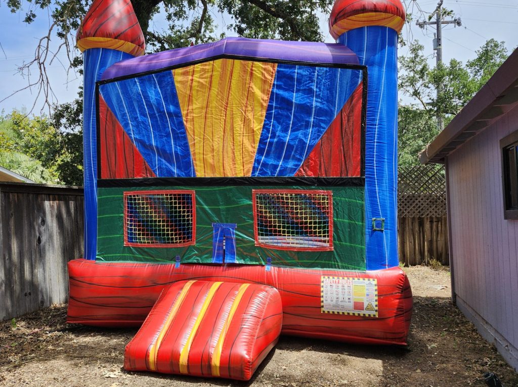 A brightly colored inflatable castle bounce house with red, blue, yellow, and green panels, set up in a backyard with trees and a wooden fence in the background.